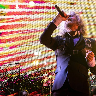Wayne Coyne singt mit angewinkelten Arm und zurückgeworfenen Kopf ins Mikro. Er trägt einen dunkelblauen Anzug vor gelb-roter Videoleinwand.