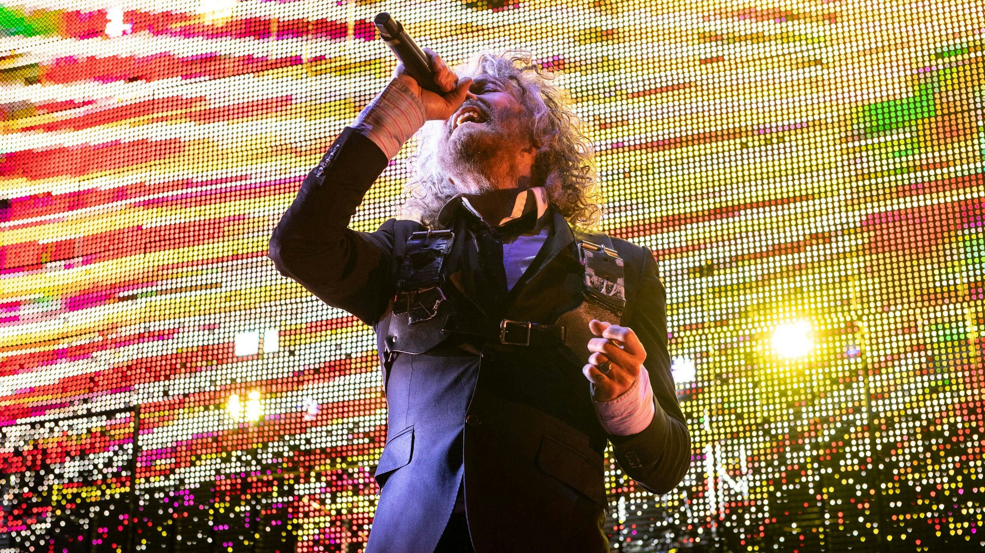 Wayne Coyne singt mit angewinkelten Arm und zurückgeworfenen Kopf ins Mikro. Er trägt einen dunkelblauen Anzug vor gelb-roter Videoleinwand.