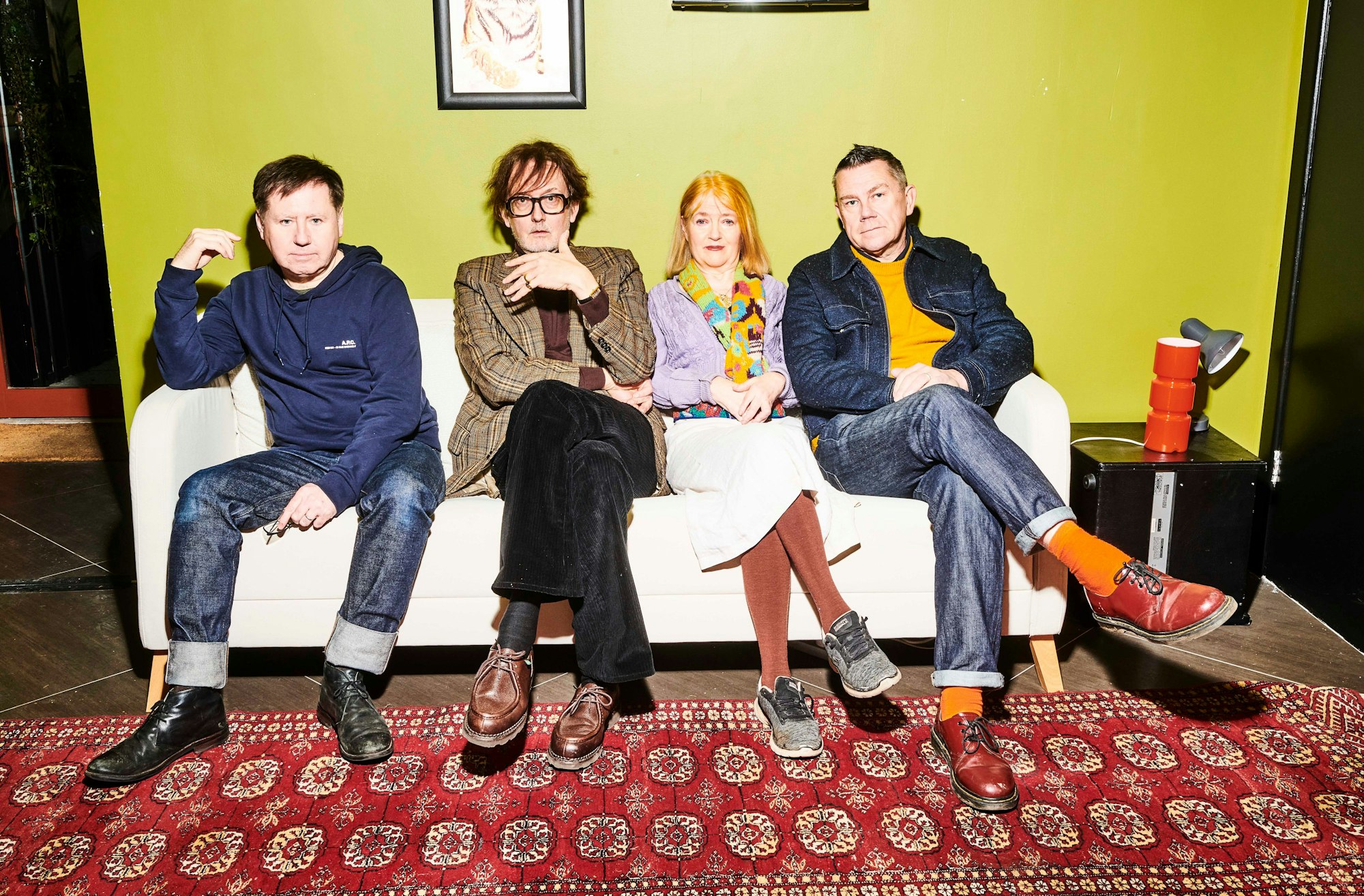 Die vier verbliebenen Mitglieder von Pulp sitzen auf einem weißen Sofa vor hellgrüner Wand.
