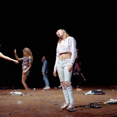 Eine Tänzerin steht mit abgewinkelten Kopf in der Mitte der torfbedeckten Bühne.