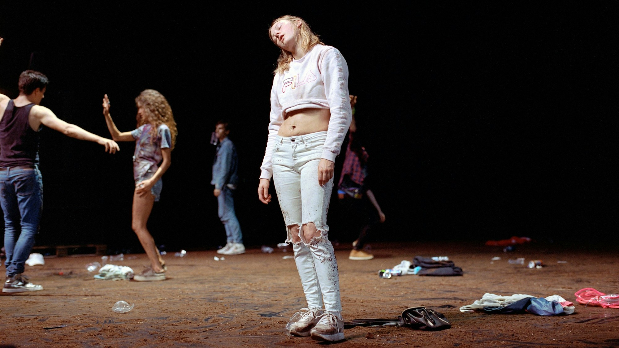 Eine Tänzerin steht mit abgewinkelten Kopf in der Mitte der torfbedeckten Bühne.