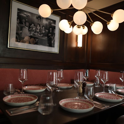 Ein eingedeckter Tisch unter einer mehrleuchtigen Lampe ist zu sehen. Über der Sitzbank auf der holzvertäfelten Wand ein Schwarzweißfoto in Rahmen.