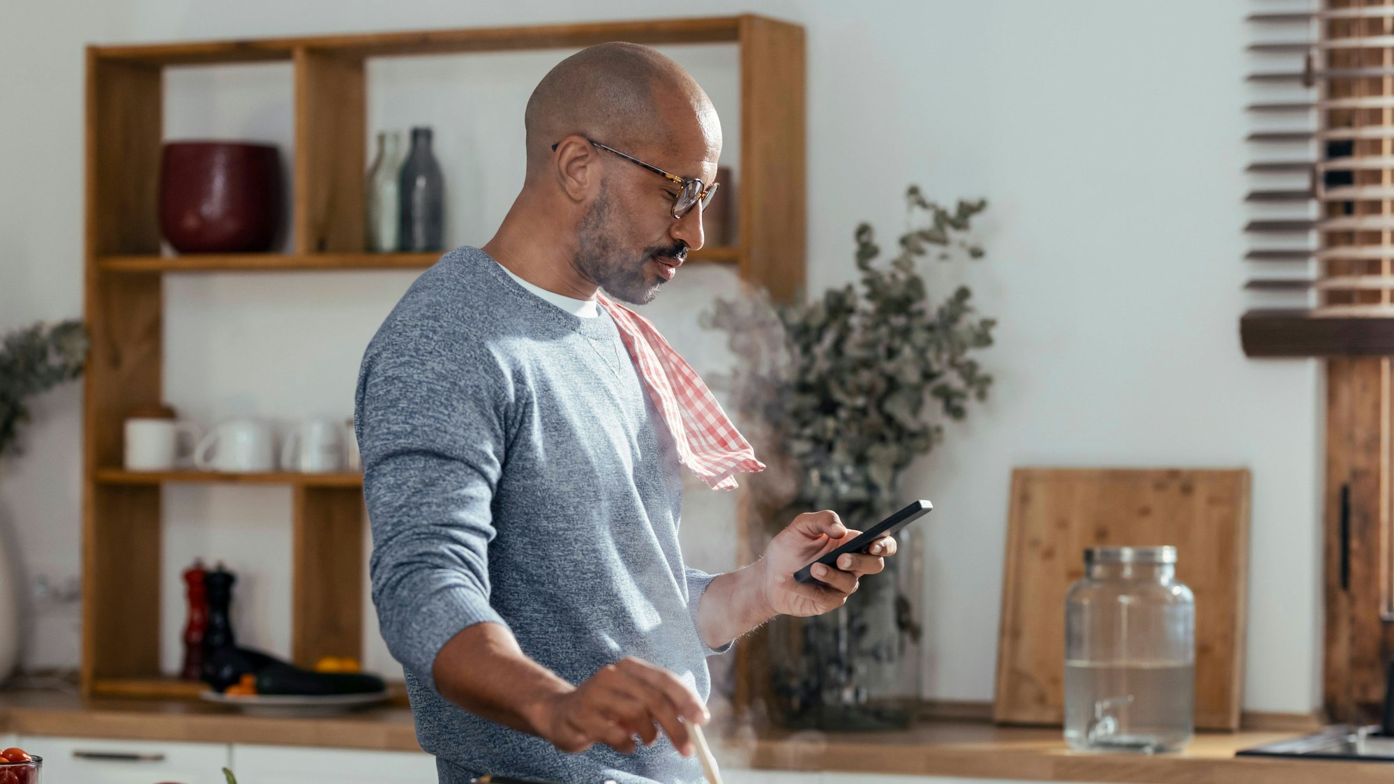 Ein Mann, der in der Küche einen Topf umrührt und sein Smartphone benutzt.