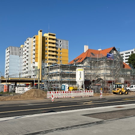 Das Bild zeigt den Bürgerbahnhof von Gerüsten umgeben, davor das bunte Treiben der Baustelle mit Verkehrsbarrieren und Fahrzeugen.