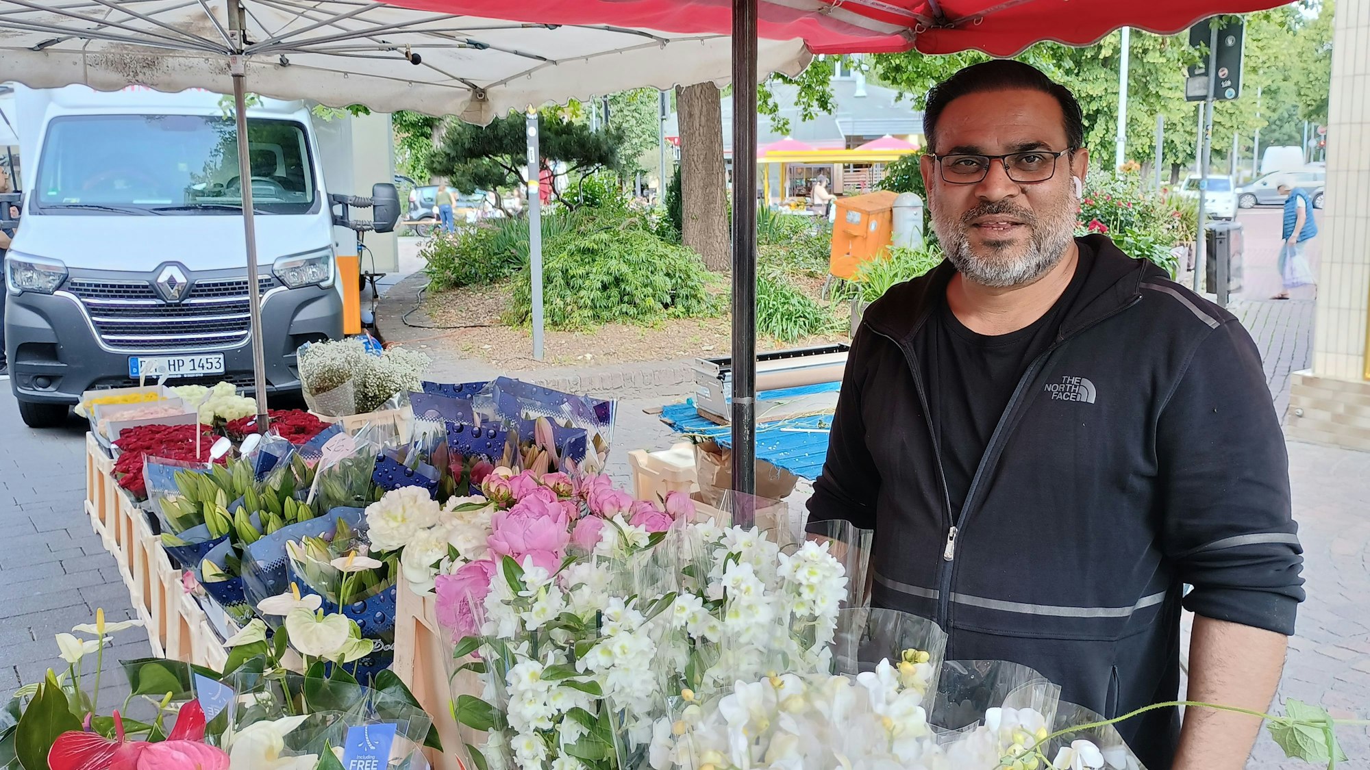 Der Blumenhändler trägt einen Pullover mit Reißverschluss. Vor ihm sind seine Blumen aufgestellt.
