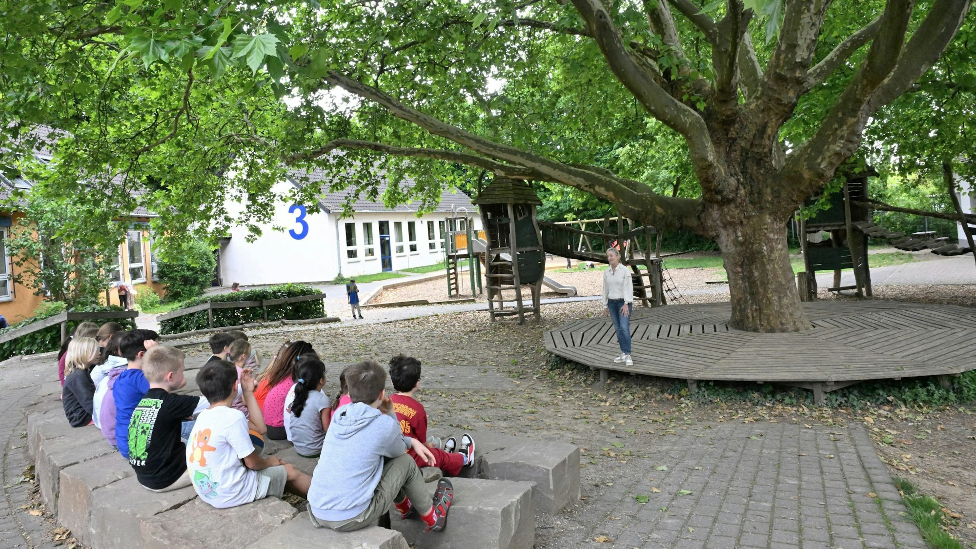 Auf Steinblöcken sitzen Kinder unter dem Blätterdach einer Platane.