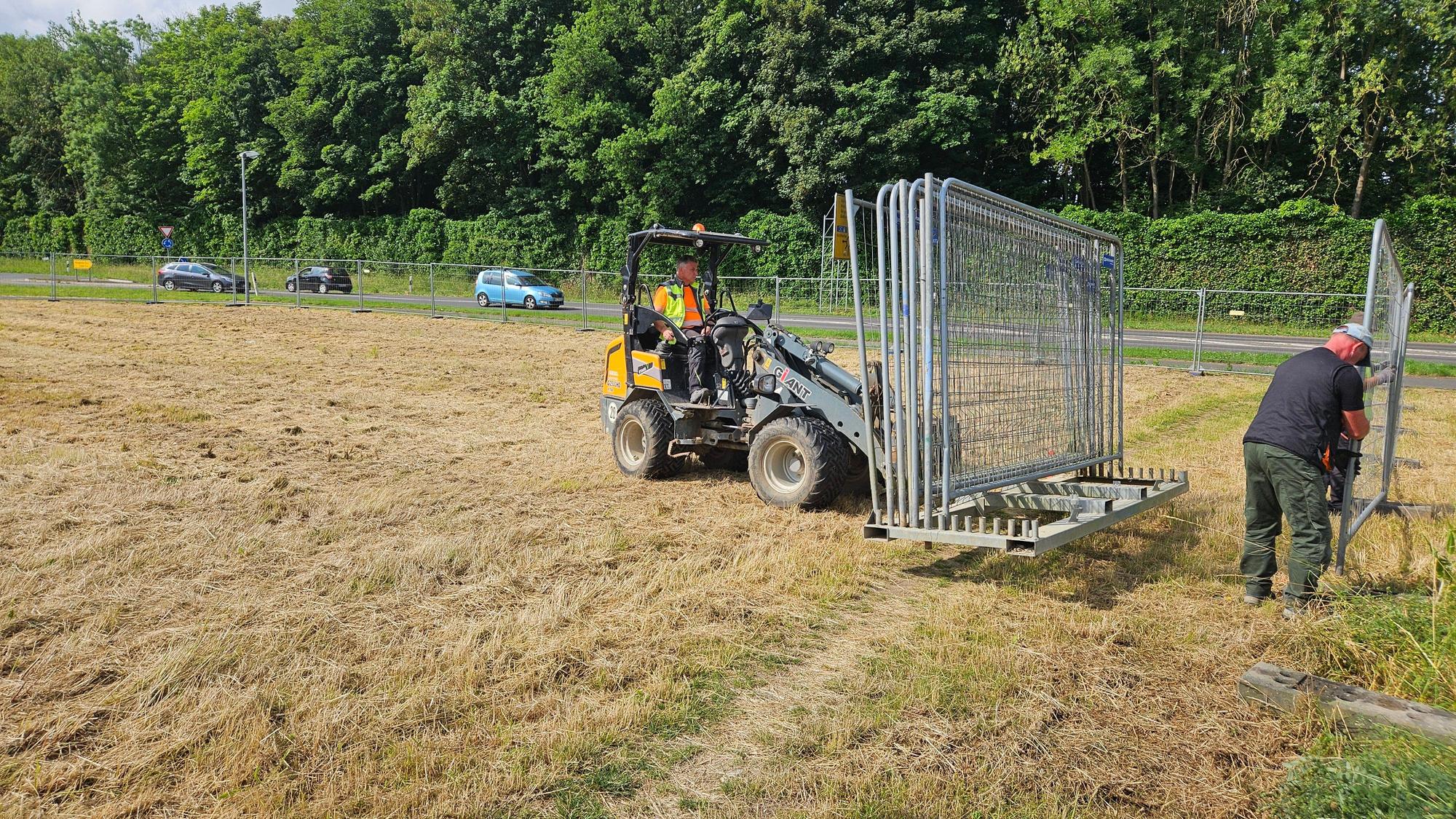 Zu sehen sind ein Minibagger auf einer Wiese und Arbeiter, die einen Bauzaun aufstellen.