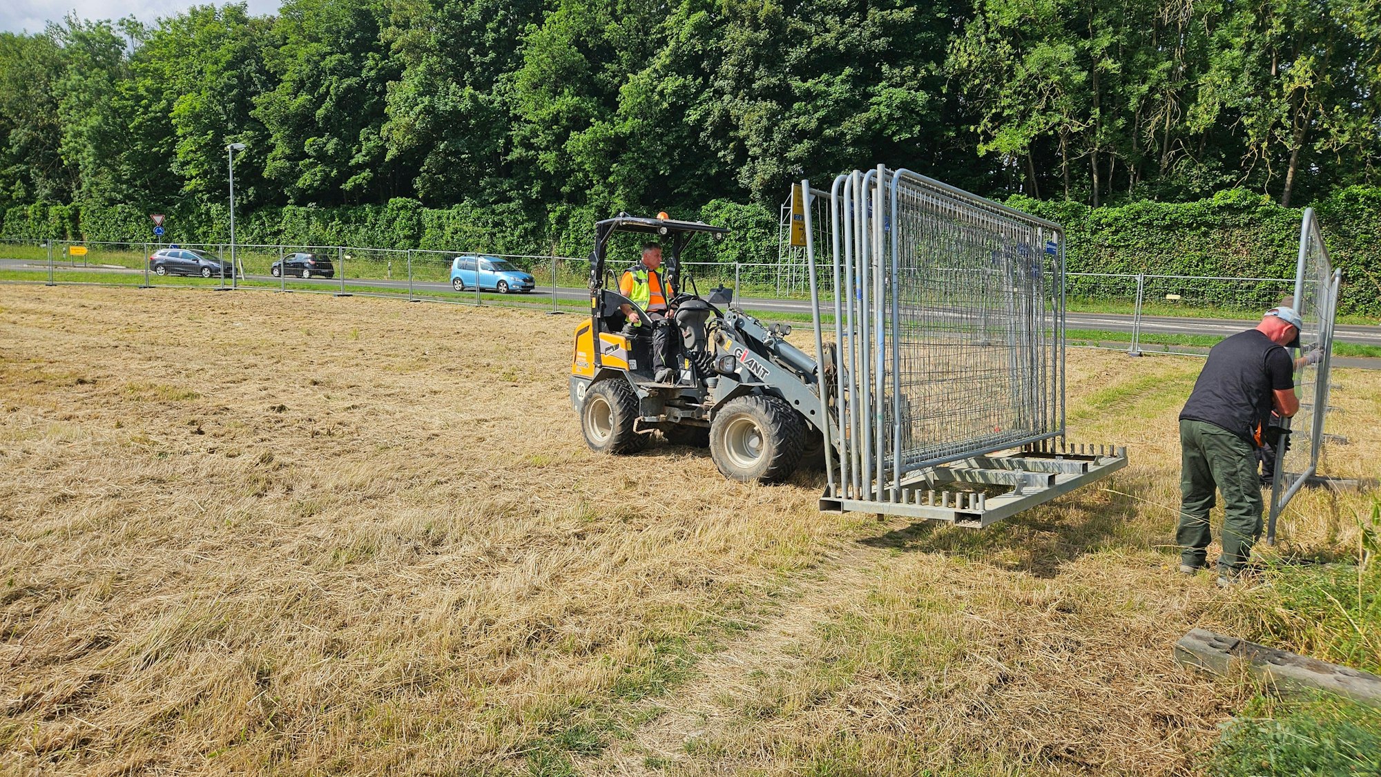Zu sehen sind ein Minibagger auf einer Wiese und Arbeiter, die einen Bauzaun aufstellen.