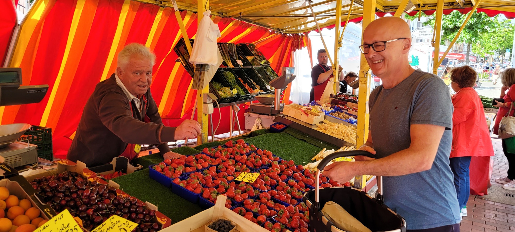 Auf dem Bild ist ein Obst- und Gemüsestand mit einem Händler und einem Kunden zu sehen.