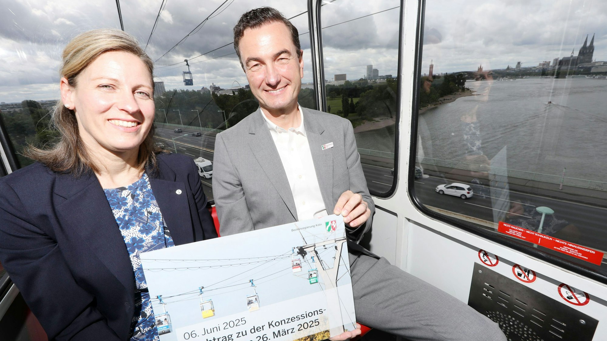 Zu sehen sind Regierungspräsident Thomas Wilk und Sonja Lorsy, Geschäftsführerin der Kölner Seilbahn, in einer Seilbahn über dem Rhein.