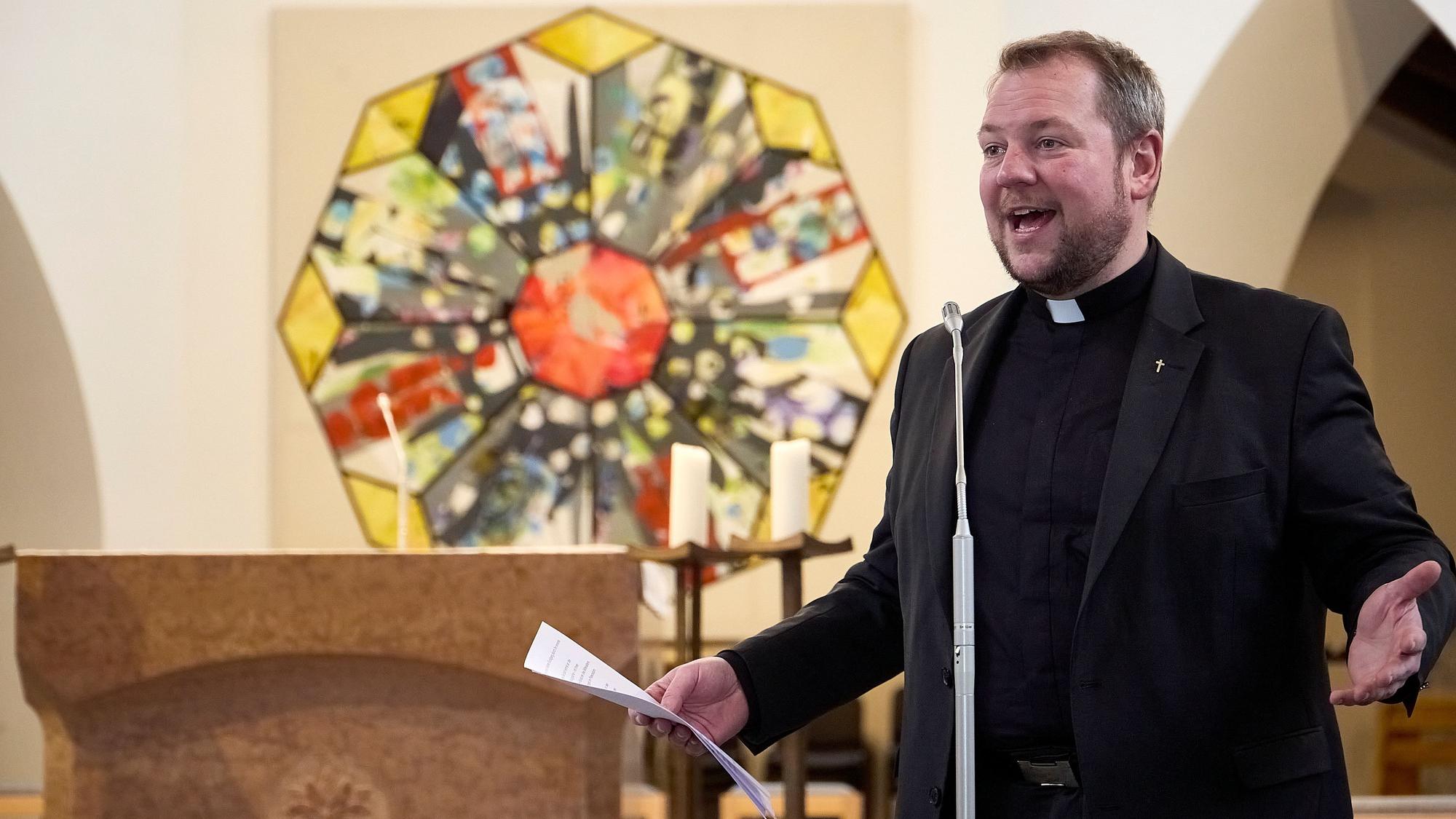 Pfarrer Thomas Schlütter steht in Schwarz gekleidet an einem Mikrofon und hält ein Blatt Papier in der Hand. Seine Arme sind ausgestreckt, sein Gesichtsausdruck ist enthusiastisch. Im Hintergrund ist ein Gemälde eines oktogonalen Kirchenfensters zu sehen.