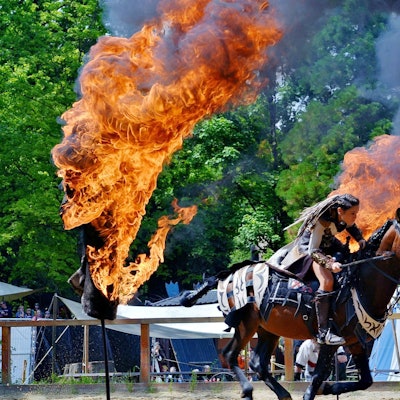 Eine Frau mit geflochtenen Haaren und Kampfausrüstung reitet zwischen zwei Feuertonnen hindurch.