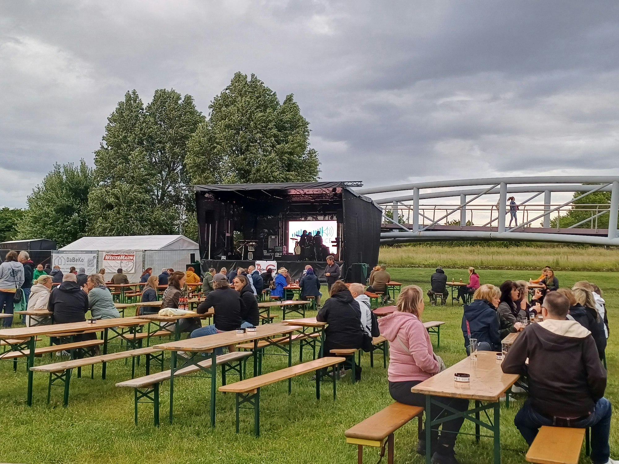 Besucher sitzen in einem Park auf Bierbänken vor einer Bühne.
