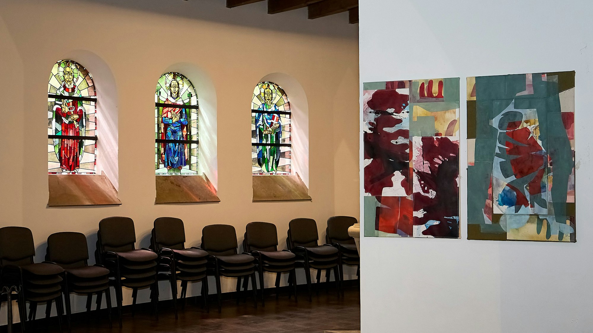 Sonne scheint durch drei bunte Kirchenfenster, an einer Säule daneben finden sich die Farben der Fenster auf Gemälden wieder.