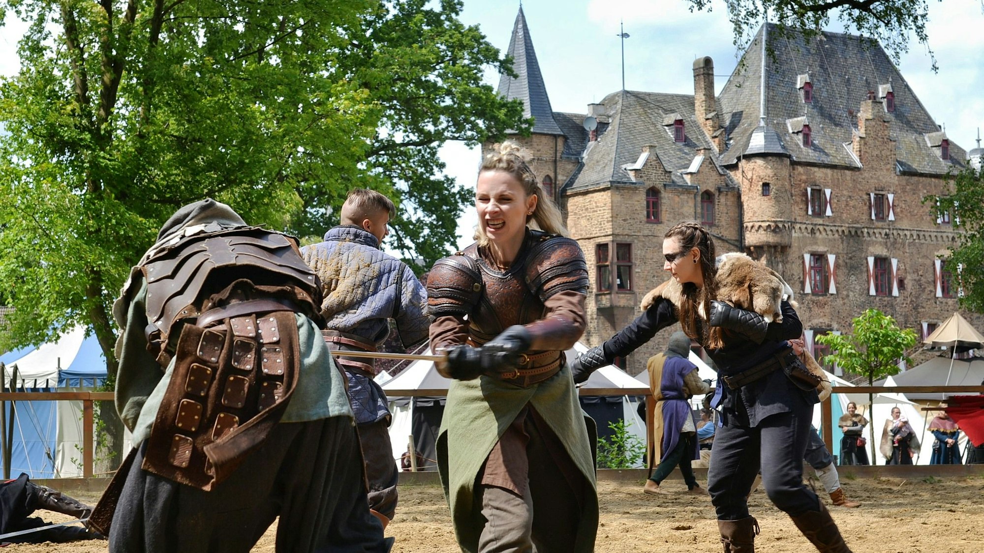 Eine Frau in mittelalterlicher Kampfausrüstung sticht mit einem Schwert einen Gegner nieder. Im Hintergrund ist die Burg Satzvey zu erkennen.