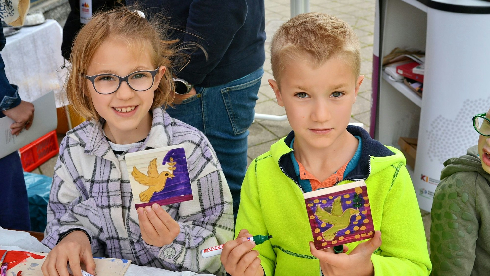 Zwei Kinder halten bemalte Holz-Vierecke in die Kamera. Darauf sind Friedenstauben abgebildet.