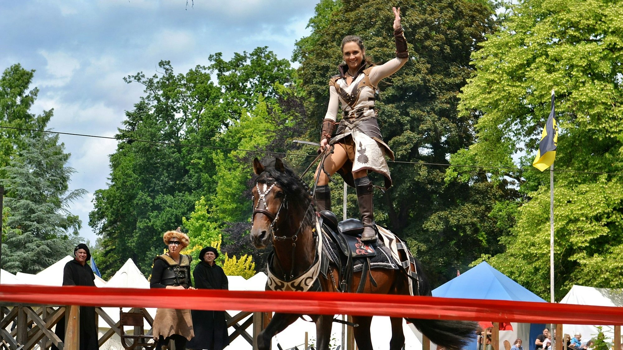 Eine Frau in mittelalterlicher Kluft reitet stehend auf einem braunen Pferd.