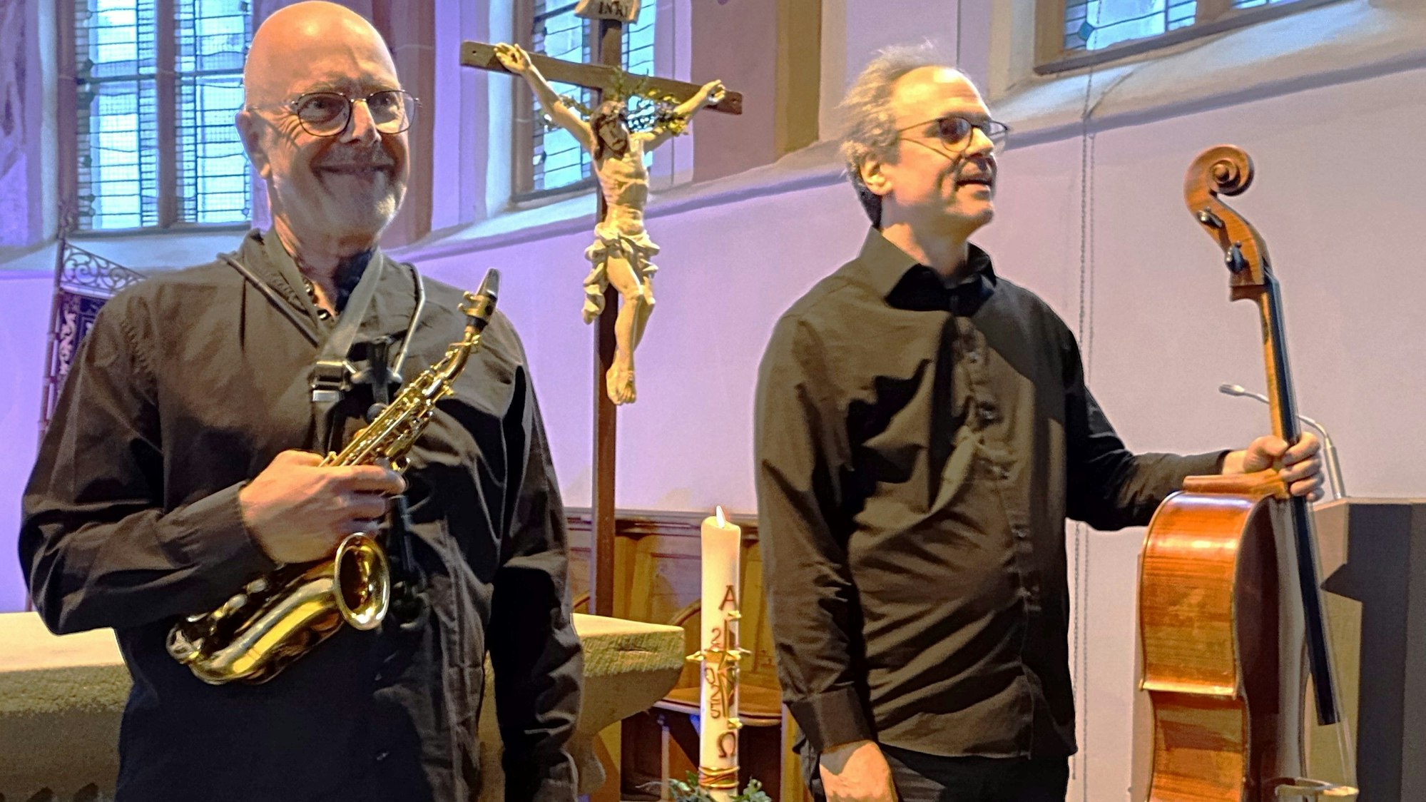 Zu sehen ist ein Musiker mit einem Saxophon und einer mit Cello vor einem Kreuz in einer Kirche.