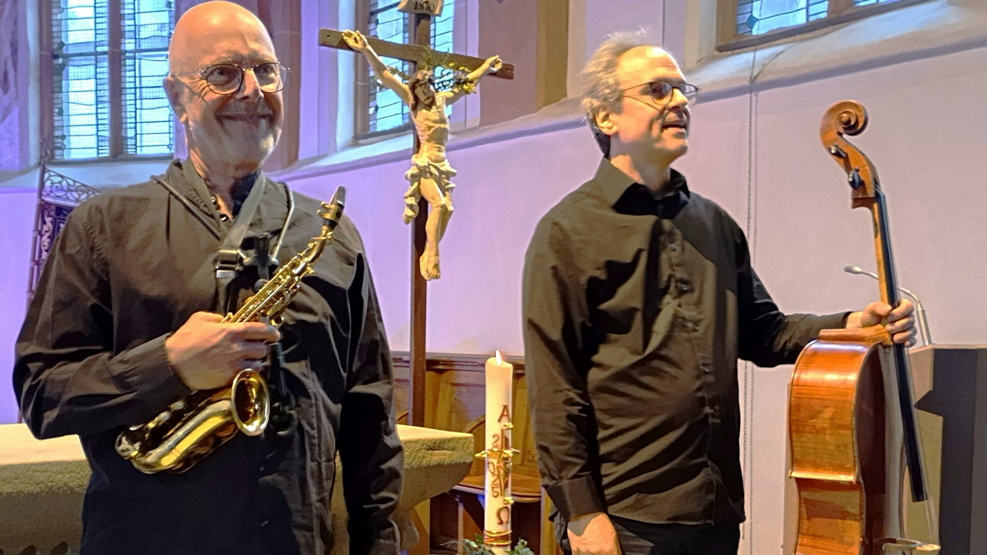 Zu sehen ist ein Musiker mit einem Saxophon und einer mit Cello vor einem Kreuz in einer Kirche.