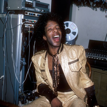 Sly Stone trägt einen goldlaminierten Anzug. Er sitzt vor einem Mischpult und hat den Mund lachend weit geöffnet.