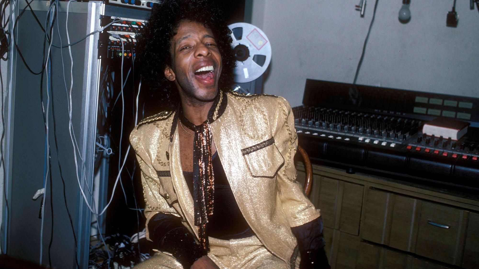 Sly Stone trägt einen goldlaminierten Anzug. Er sitzt vor einem Mischpult und hat den Mund lachend weit geöffnet.