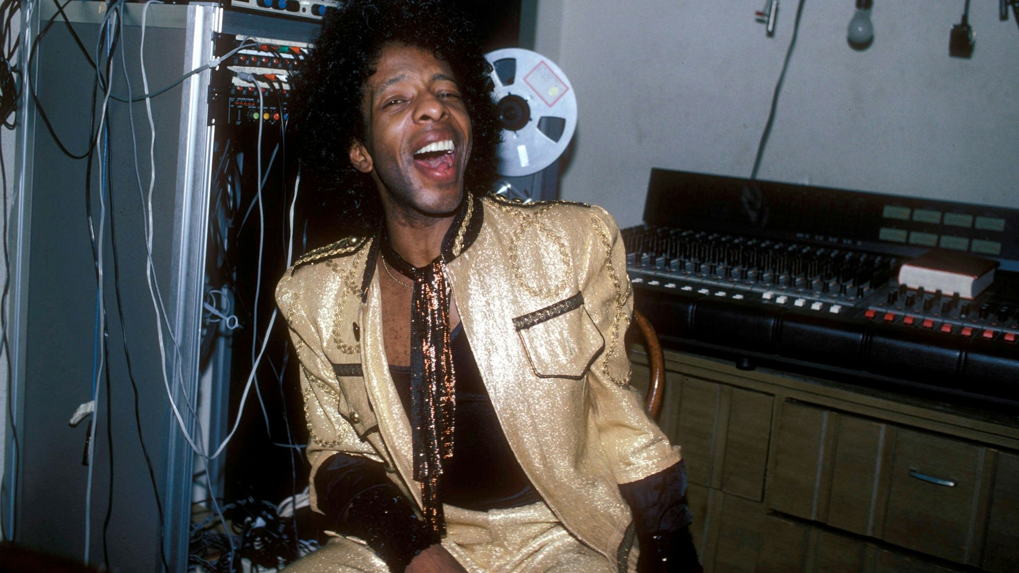 Sly Stone trägt einen goldlaminierten Anzug. Er sitzt vor einem Mischpult und hat den Mund lachend weit geöffnet.