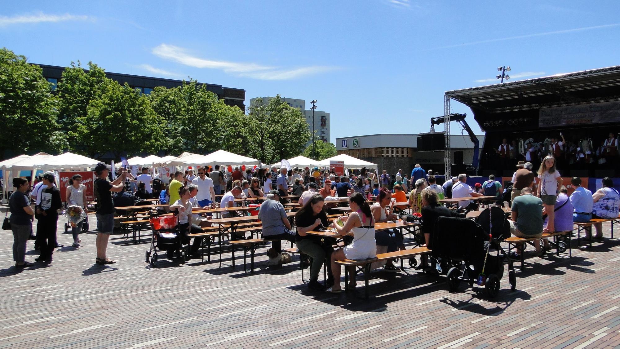 Zu sehen sind Menschen, die auf Biergarnituren sitzen, in der Sonne auf dem Pariser Platz in Chorweiler. Links im Bild sind weiße Pavillons und grüne Bäume zu sehen, recht eine Bühne.