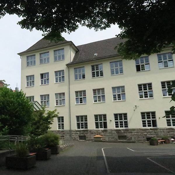 Ein mehrgeschossiges herkömmliches Schulgebäude mit vorgelagertem Schulhof und Grünanlage ist zu sehen.