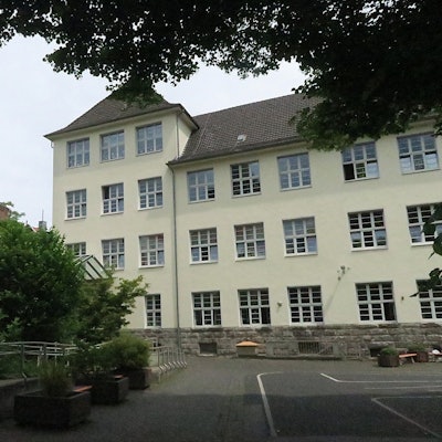 Ein mehrgeschossiges herkömmliches Schulgebäude mit vorgelagertem Schulhof und Grünanlage ist zu sehen.