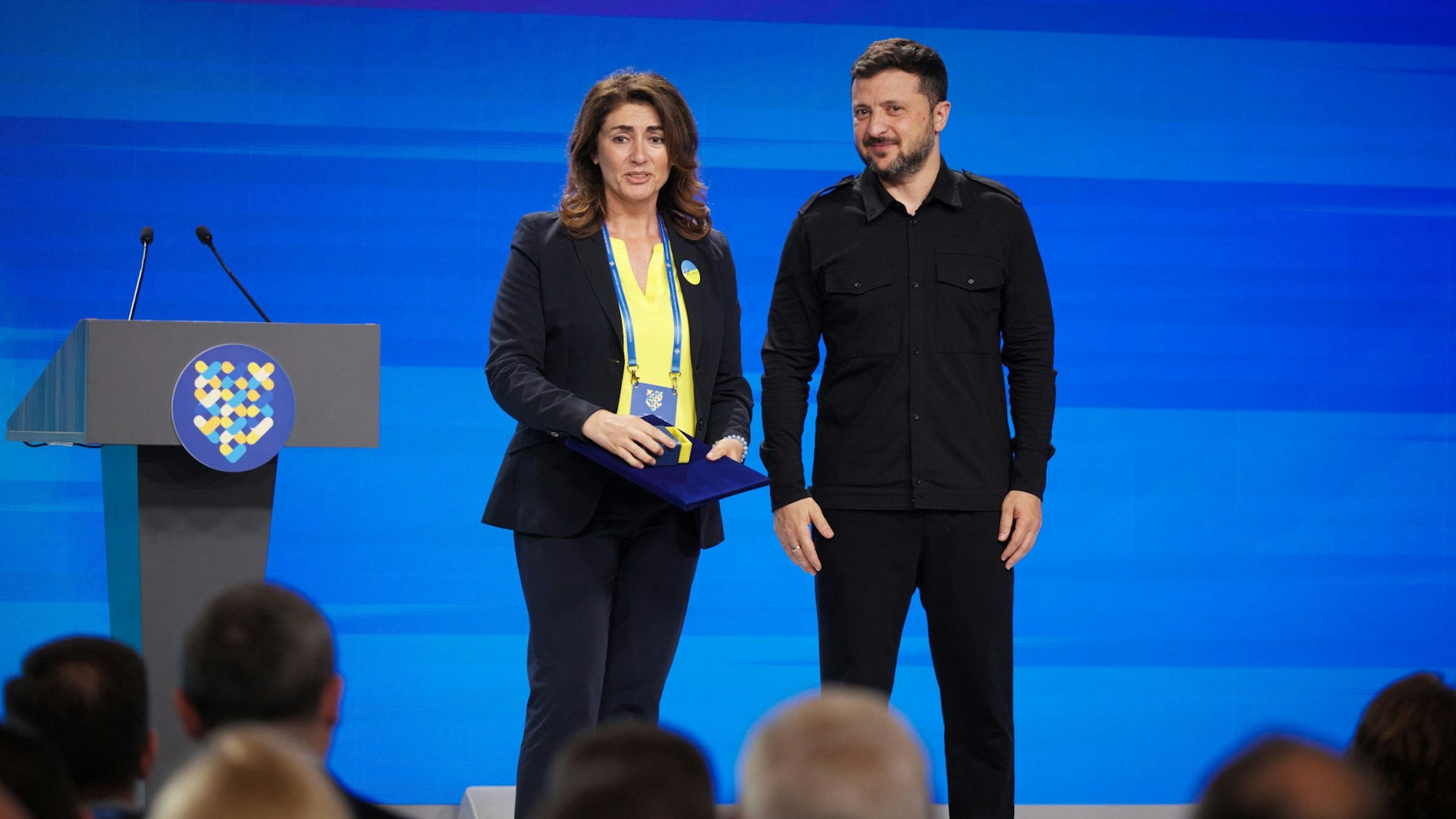 Wolodymyr Selenskyj steht neben Linda Mai auf einer Bühne.