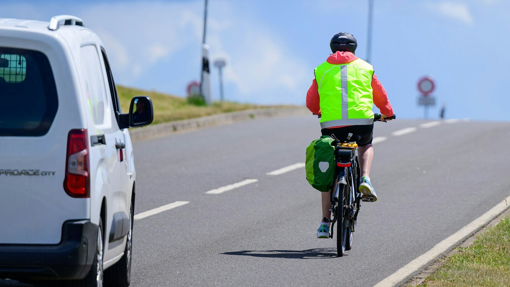 Ein Radfahrer mit einer gelben Weste fährt auf der Straße, ein Auto fährt dahinter.