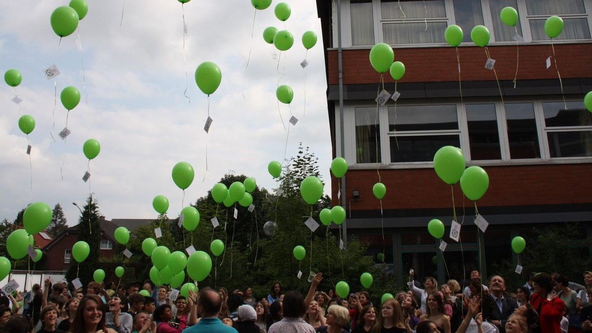 Viele Schüler und Eltern stehen auf dem Schulhof und lassen grüne Luftballons in den Himmel steigen.