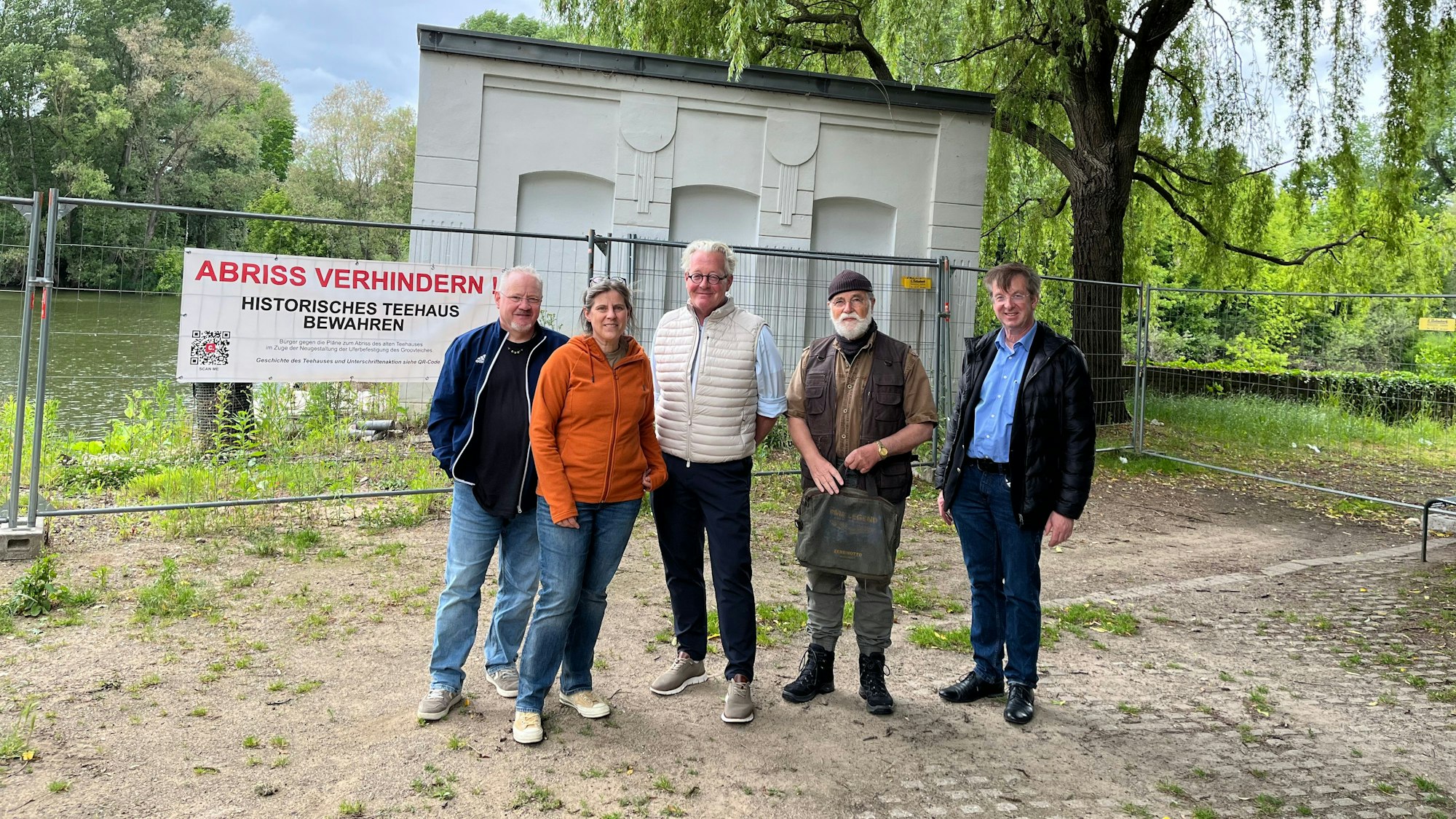 Fünf Menschen stehen vor einem kleinen Gebäude, das hinter einem Bauzaun steht.