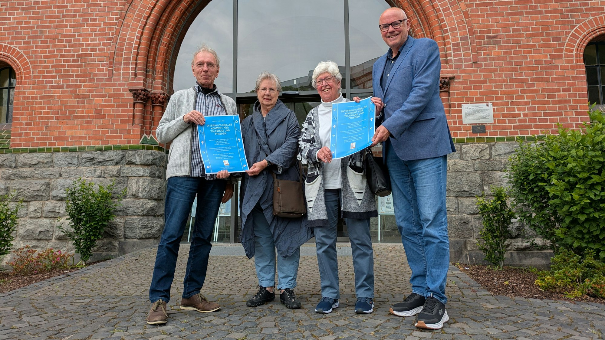 Zwei Männer und zwei Frauen stehen vor einer Kirche und halten Plakate hoch.