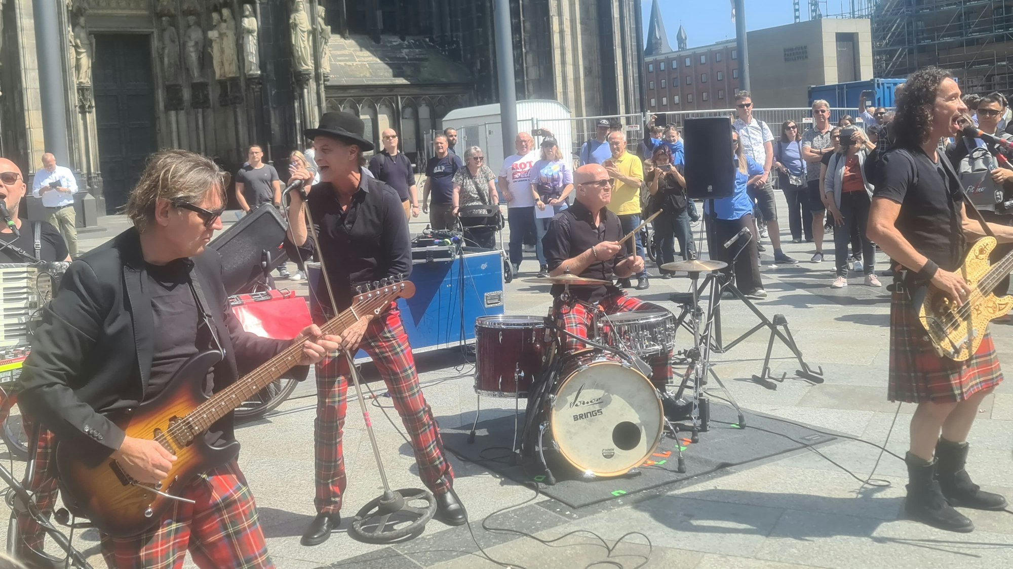 Zu sehen ist die Band Brings, die ein Konzert auf der Domplatte spielen. Im Hintergrund sind der Kölner Dom, sowie Zuschauerinnen und Zuschauer zu sehen.
