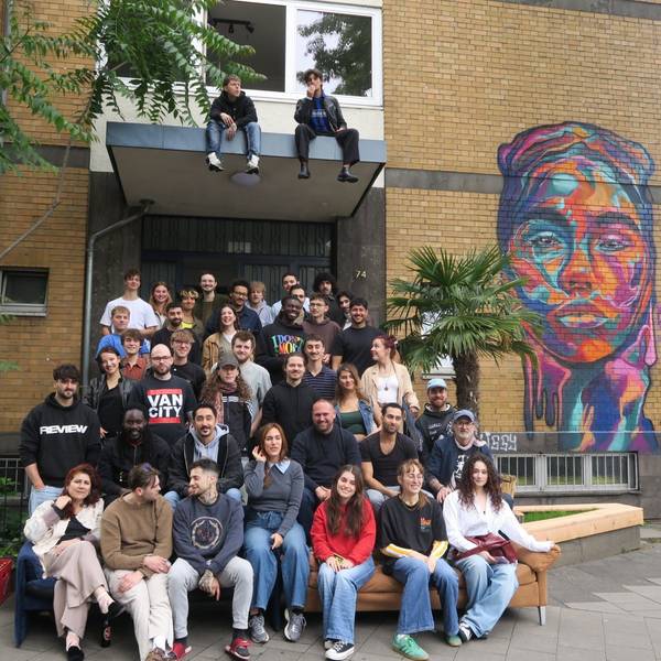 Eine große Gruppe junger Menschen sitzt vor einem Gebäude, auf das ein buntes Kopf-Graffito gesprüht ist.