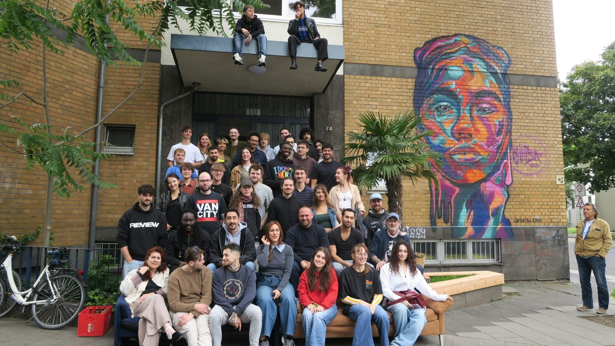 Eine große Gruppe junger Menschen sitzt vor einem Gebäude, auf das ein buntes Kopf-Graffito gesprüht ist.