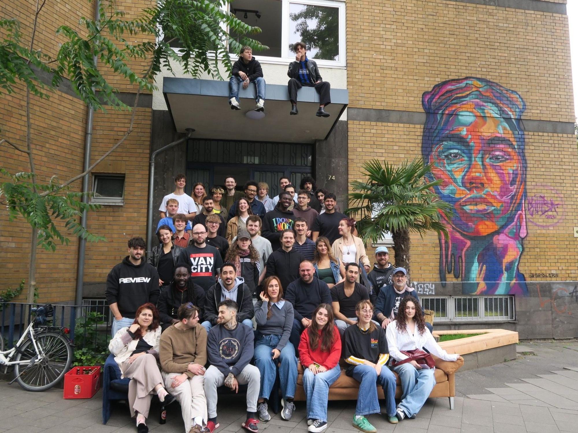 Viele junge Menschen sitzen auf den Eingangsstufen vor ihrem Wohngebäude. Sie leben in einer riesigen WG zusammen in Ehrenfeld.