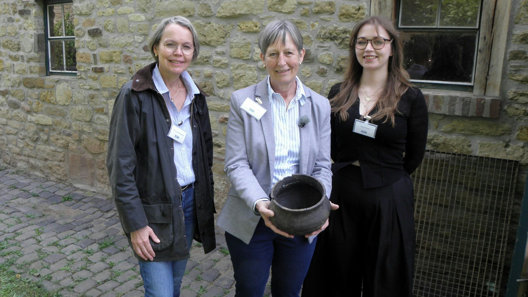 Drei Frauen stehen nebeneinander vor einer Bruchsteinfassade, Petra Tutlies in der Mitte hält ein Gefäß in den Händen.