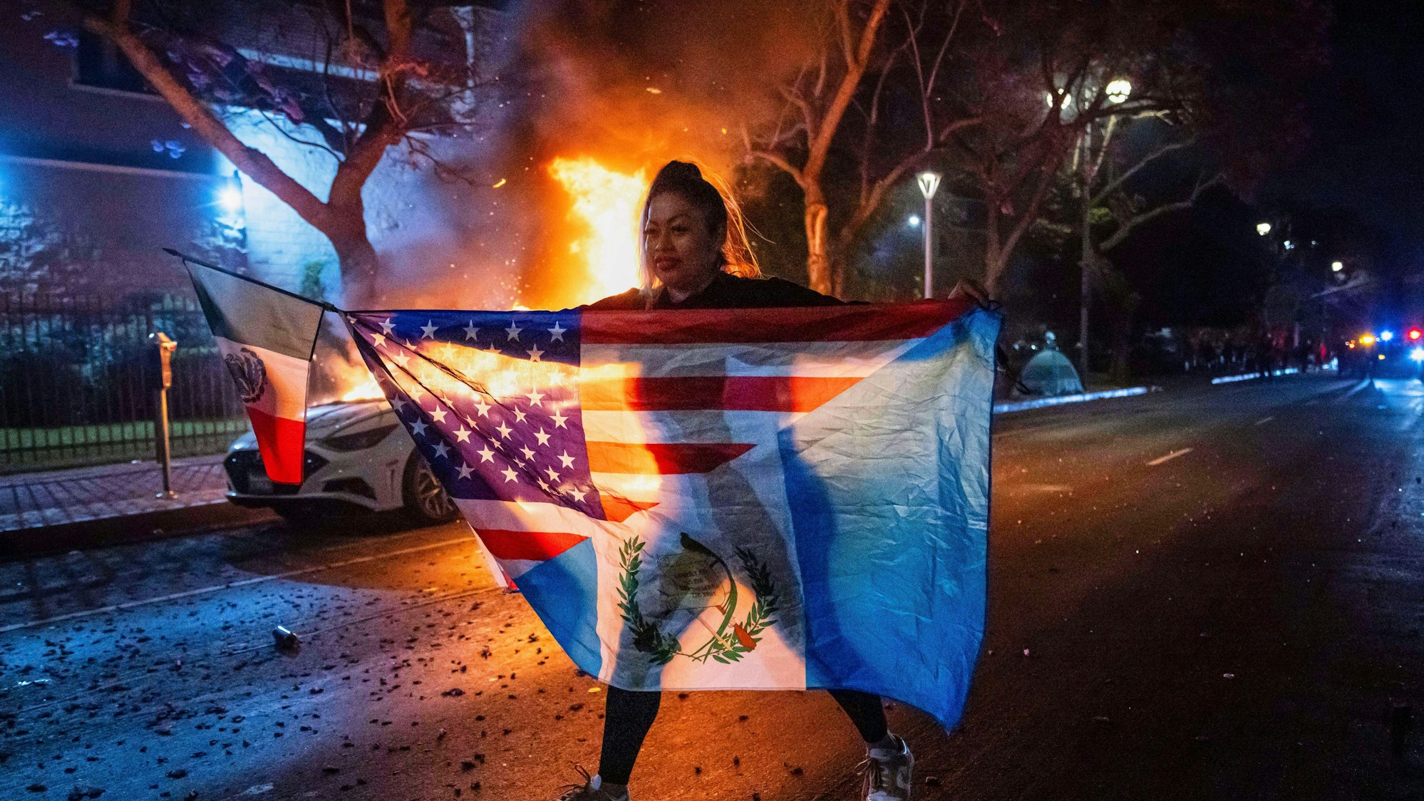 Das Bild zeigt eine Frau mit mehreren Flaggen, die an einem brennenden Auto vorbei geht, während sie gegen die Razzien der Trump-Regierung gegen Einwanderer in Los Angeles protestiert. Foto: Ethan Swope/FR171736 AP/dpa