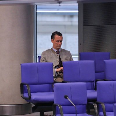 Matthias Helferich sitzt auf einem blauen Stuhl der AfD-Fraktion im Bundestag. Die Stühle um ihn herum sind leer.