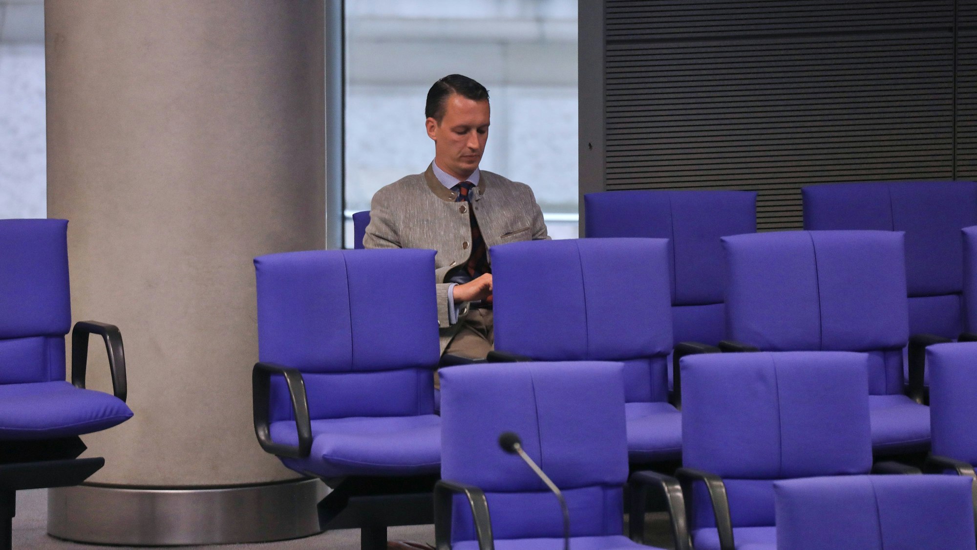 Matthias Helferich sitzt auf einem blauen Stuhl der AfD-Fraktion im Bundestag. Die Stühle um ihn herum sind leer.