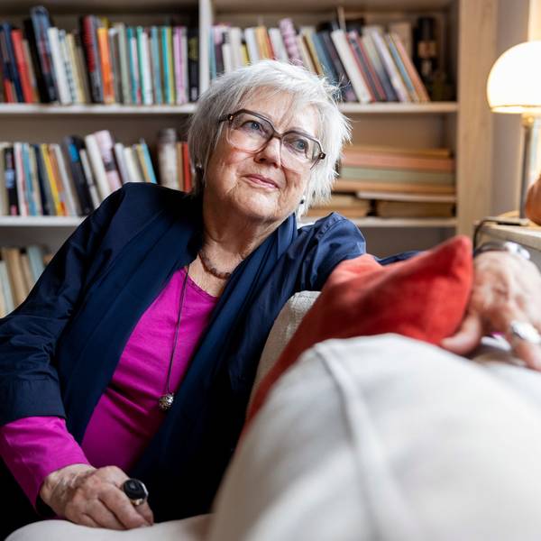 Renate Liesmann-Baum sitzt auf einer Couch in ihrem Wohnzimmer. Im Hintergrund steht ein volles Bücherregal.