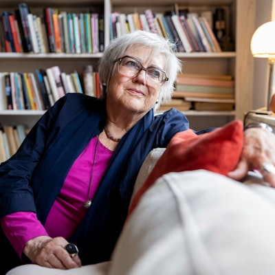 Renate Liesmann-Baum sitzt auf einer Couch in ihrem Wohnzimmer. Im Hintergrund steht ein volles Bücherregal.