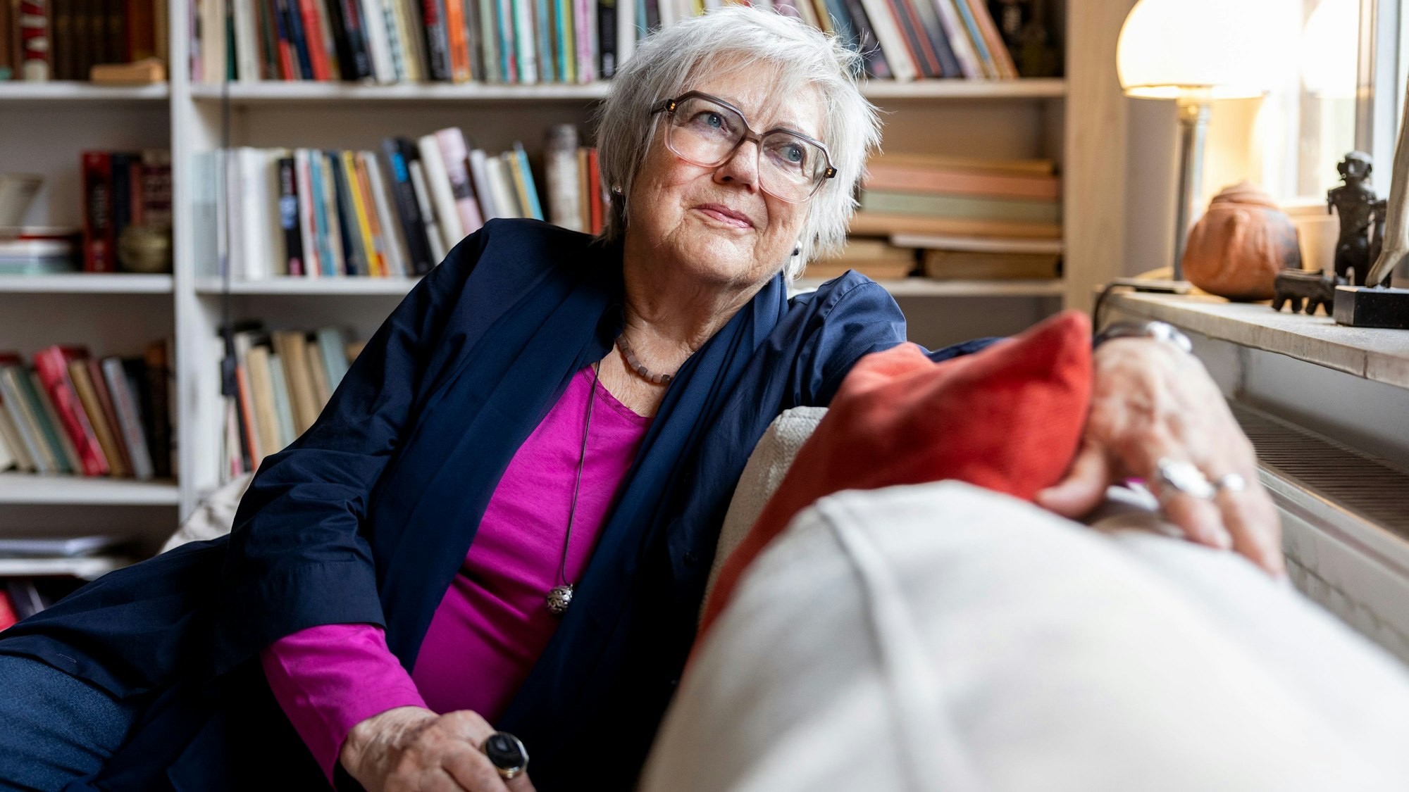 Renate Liesmann-Baum sitzt auf einer Couch in ihrem Wohnzimmer. Im Hintergrund steht ein volles Bücherregal.