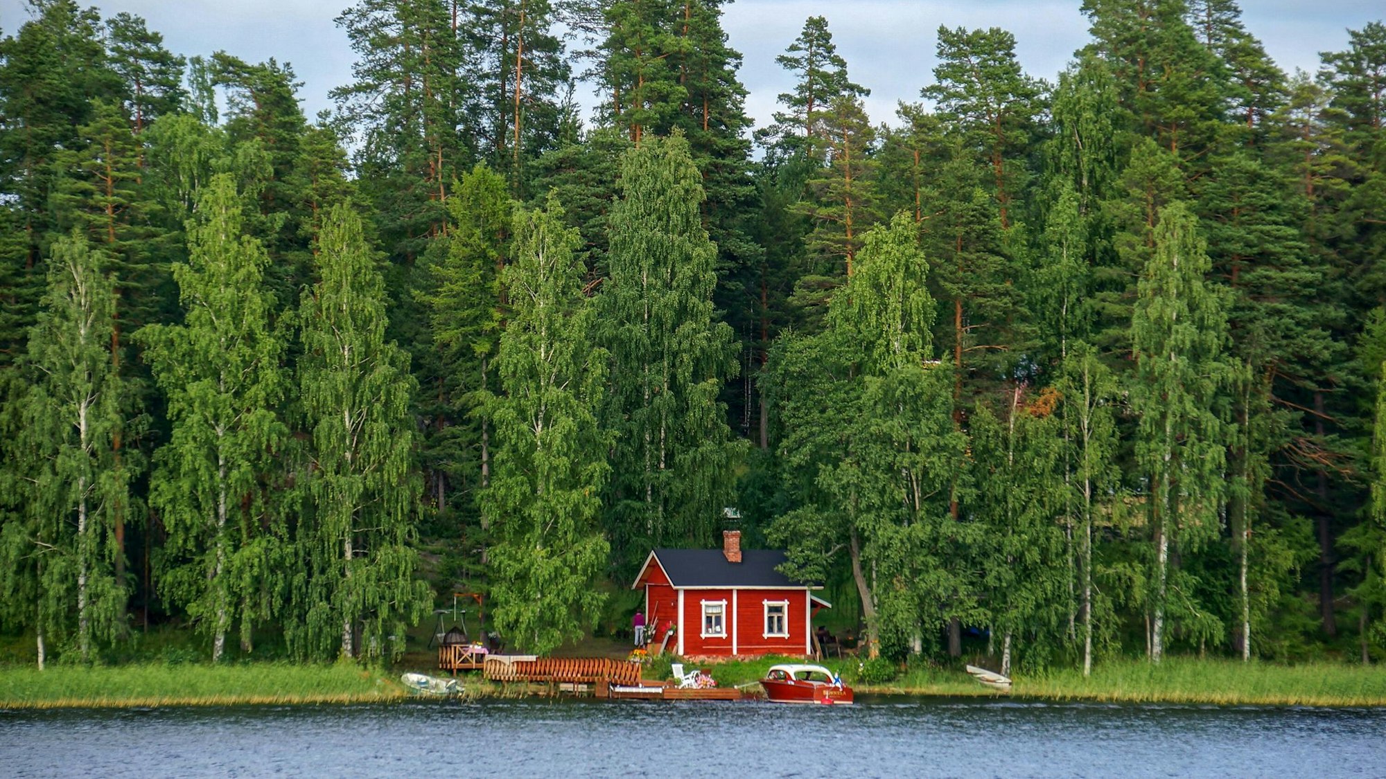 Ein kleines Häuschen an einem See, davor ein Boot, dahinter riesige Bäume.