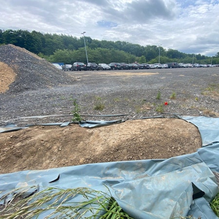 Zu sehen ist ein kleiner Erdwall, der teils mit Kunststofffolie bedeckt ist und im Hintergrund ein Parkplatz.