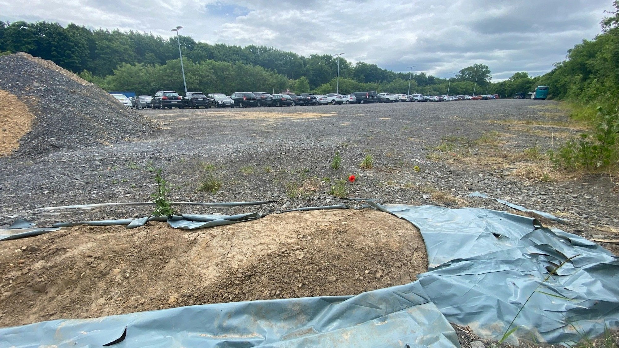 Zu sehen ist ein kleiner Erdwall, der teils mit Kunststofffolie bedeckt ist und im Hintergrund ein Parkplatz.