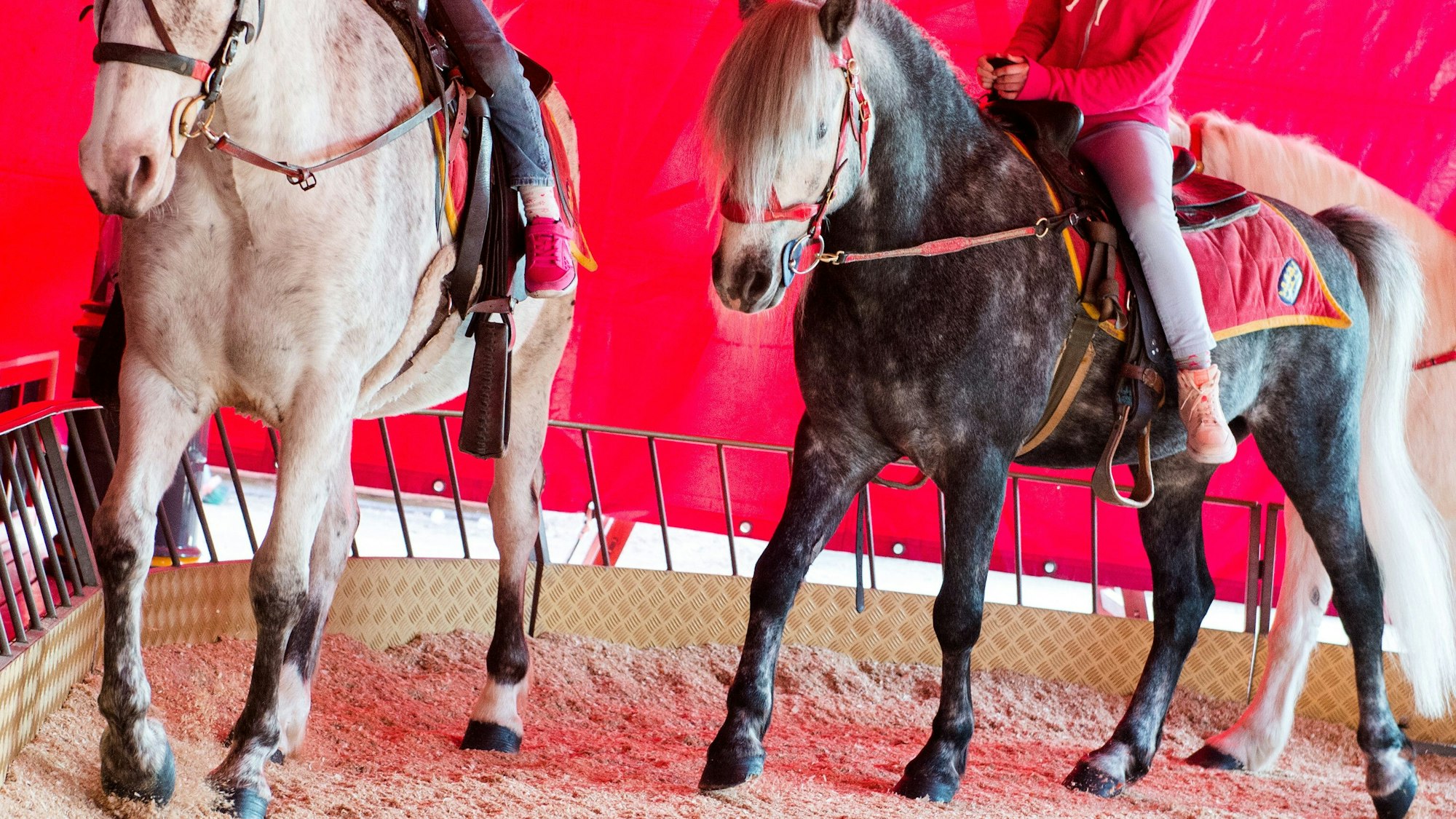 Zwei Kinder sitzen auf zwei Pferden und reiten unter einem roten Zeltdach über eine Sandstrecke.