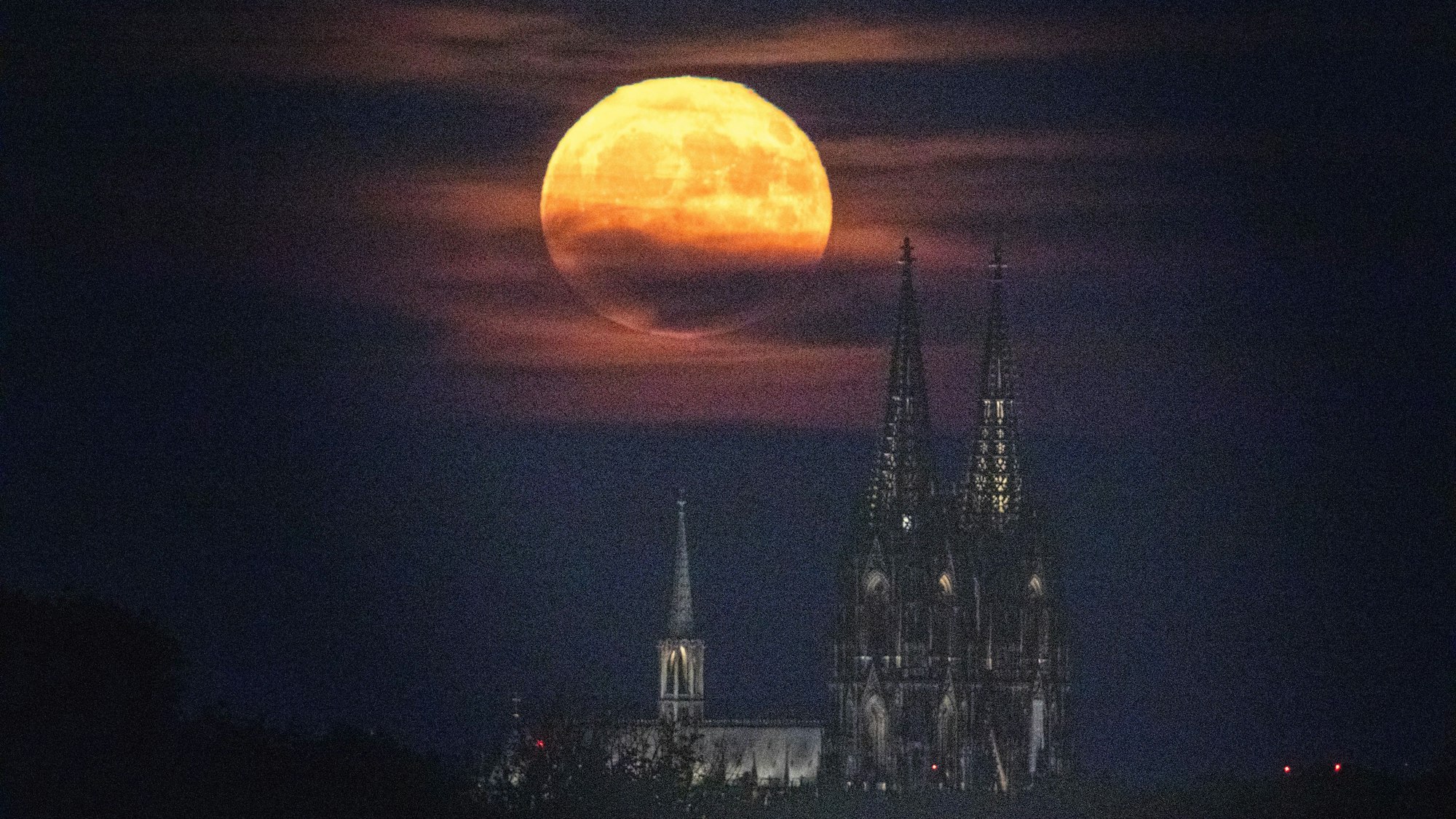 Ein gelblich-rötlicher Vollmond steht über dem Kölner Dom. Das seltene Phänomen wird auf „Erdbeermond“ genannt.